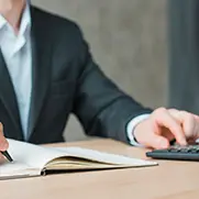 A person in a suit typing on a calculator with their left hand and writing in a notebook with their right hand.